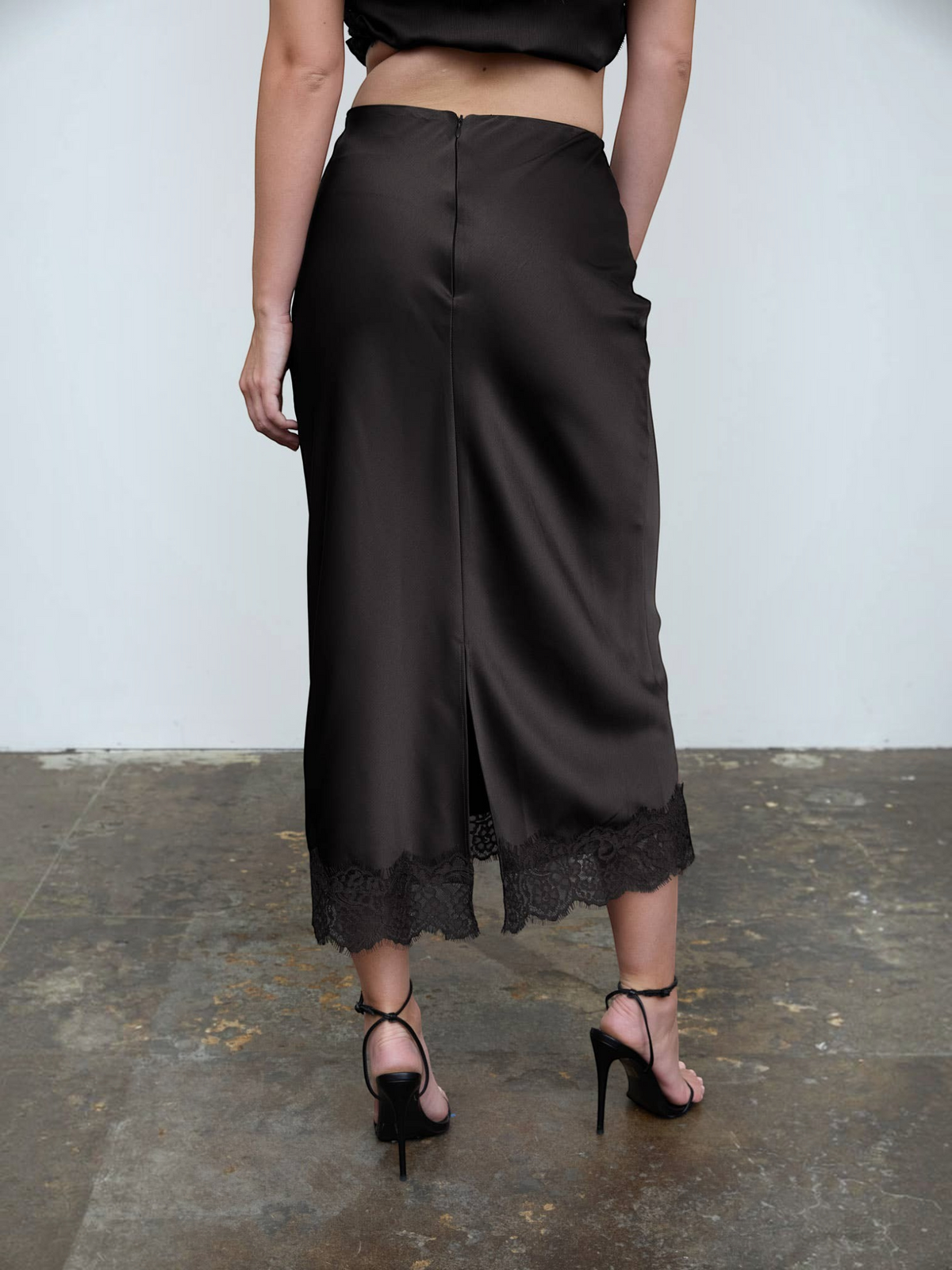 Mialur black satin midi skirt featuring a delicate lace hem and back slit detail worn with black high-heeled sandals.