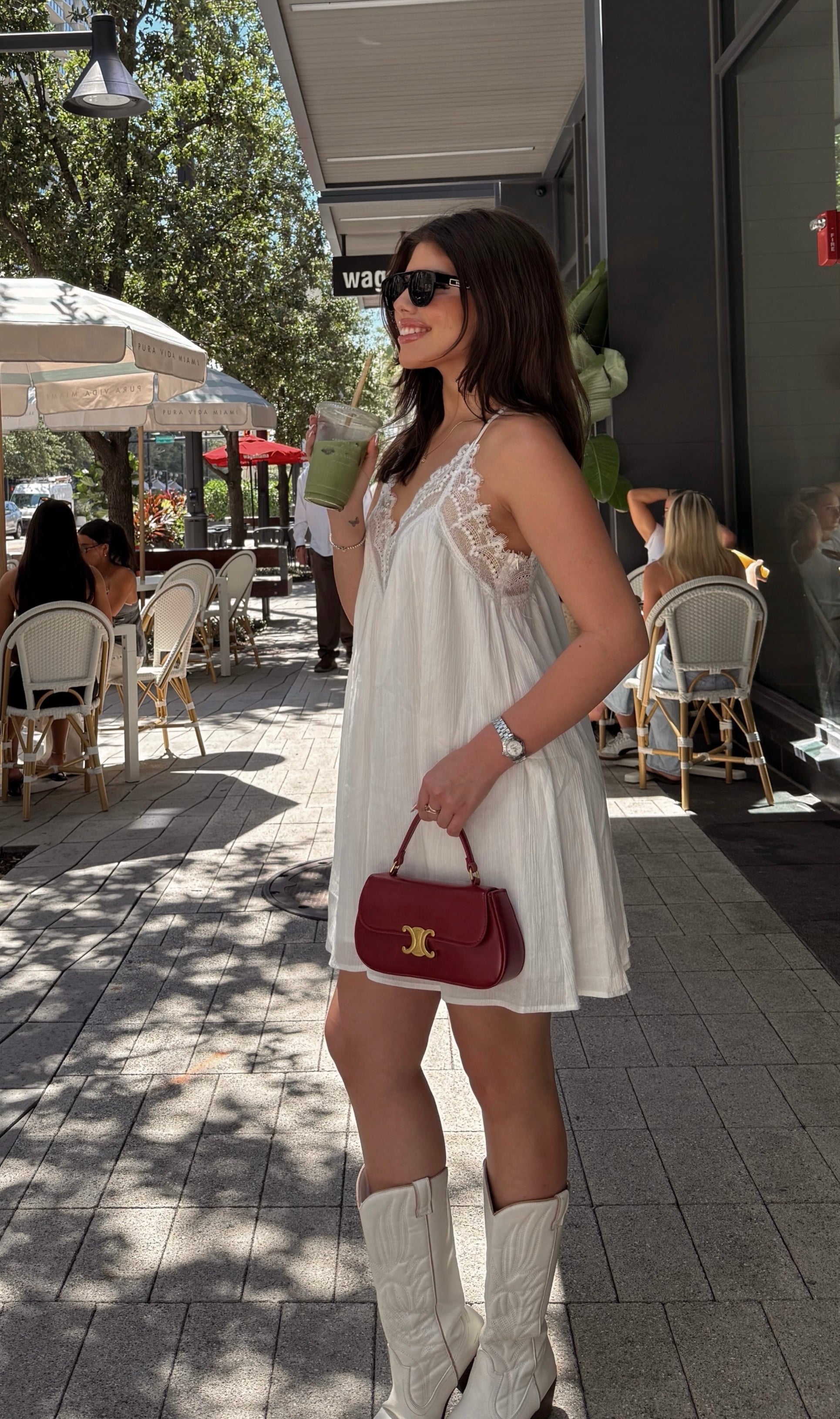 A woman wearing a white lace-trimmed sleeveless dress and white cowboy boots stands on a paved outdoor patio, holding a burgundy handbag with gold hardware.