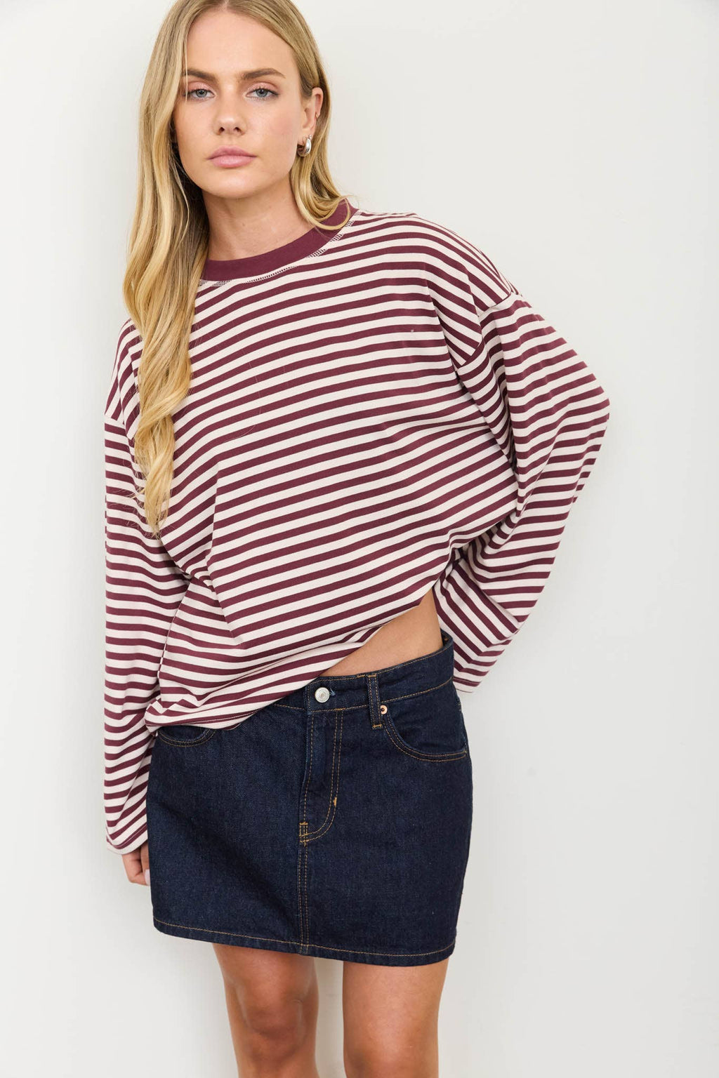 A woman modeling a long-sleeve crewneck top with horizontal burgundy and white stripes, paired with a dark denim mini skirt, against a plain light gray background.