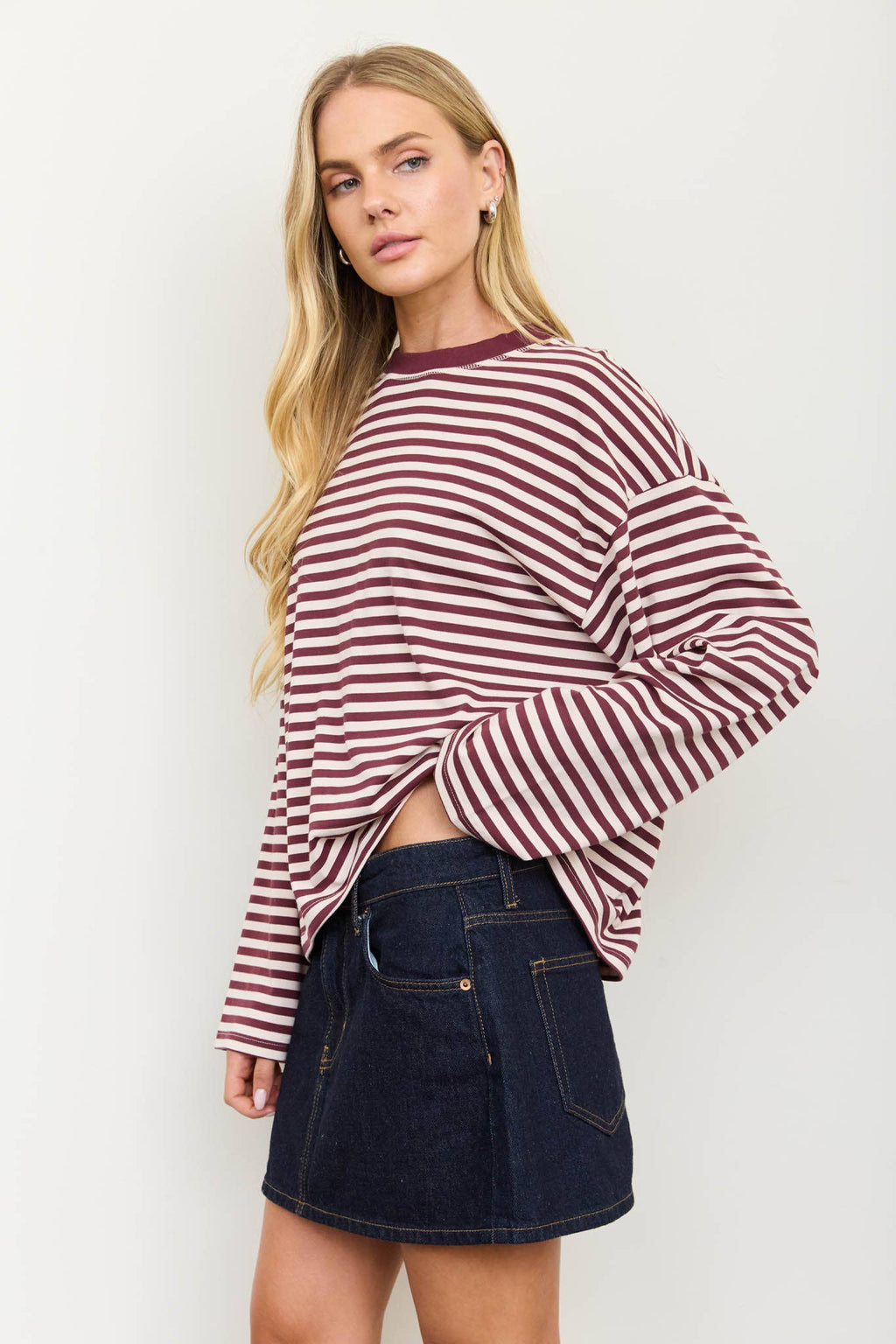 A woman modeling a long-sleeve crewneck top with horizontal burgundy and white stripes, paired with a dark denim mini skirt, against a plain off-white background.