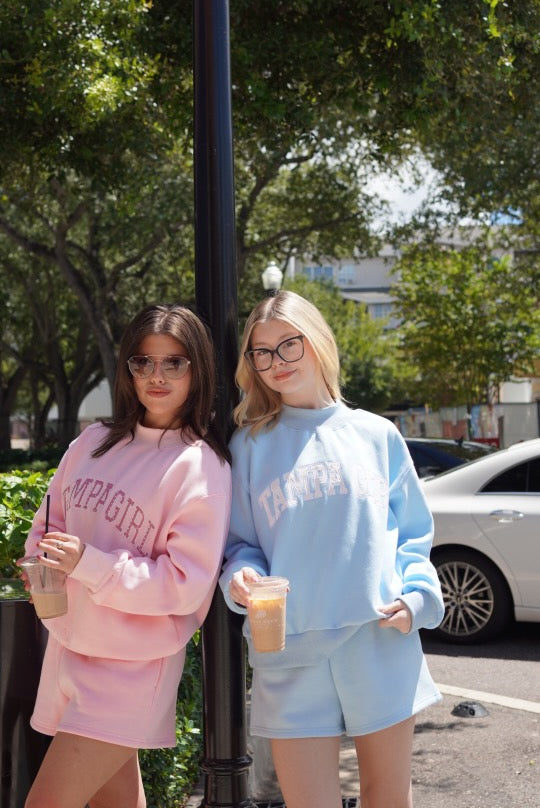 Two young women standing outdoors next to a black pole, wearing matching sweatshirt and shorts sets. The woman on the left wears a pink set with 'COFFEE GIRL' printed in dark pink on the sweatshirt and shorts.