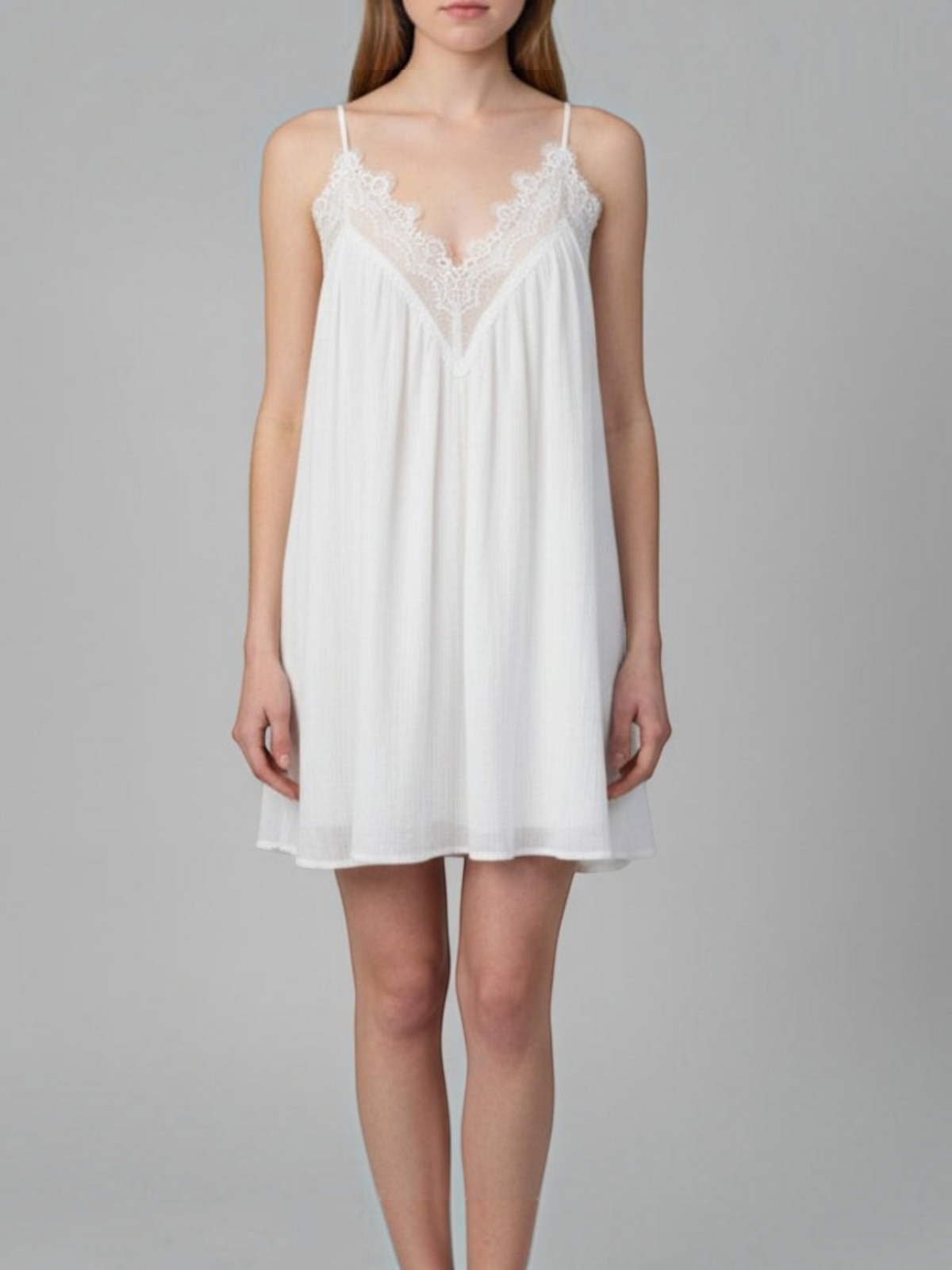 A woman modeling a white slip-style nightgown with thin straps and a lace V-neckline, photographed against a plain light gray studio background.