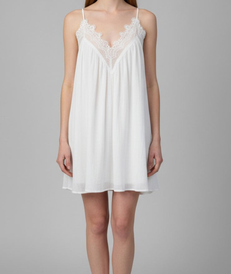 A woman modeling a white slip-style nightgown with thin straps and a lace V-neckline, photographed against a plain light gray studio background.