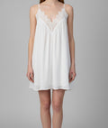 A woman modeling a white slip-style nightgown with thin straps and a lace V-neckline, photographed against a plain light gray studio background.