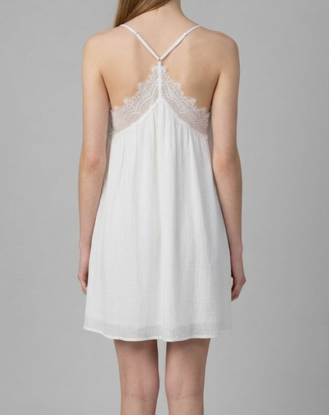 A woman shown from the back wearing a white sleeveless slip dress with thin straps and a lace-trimmed open back, photographed against a plain light gray studio background.