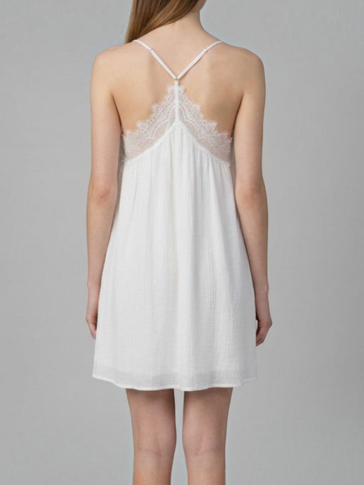 A woman shown from the back wearing a white sleeveless slip dress with thin straps and a lace-trimmed open back, photographed against a plain light gray studio background.