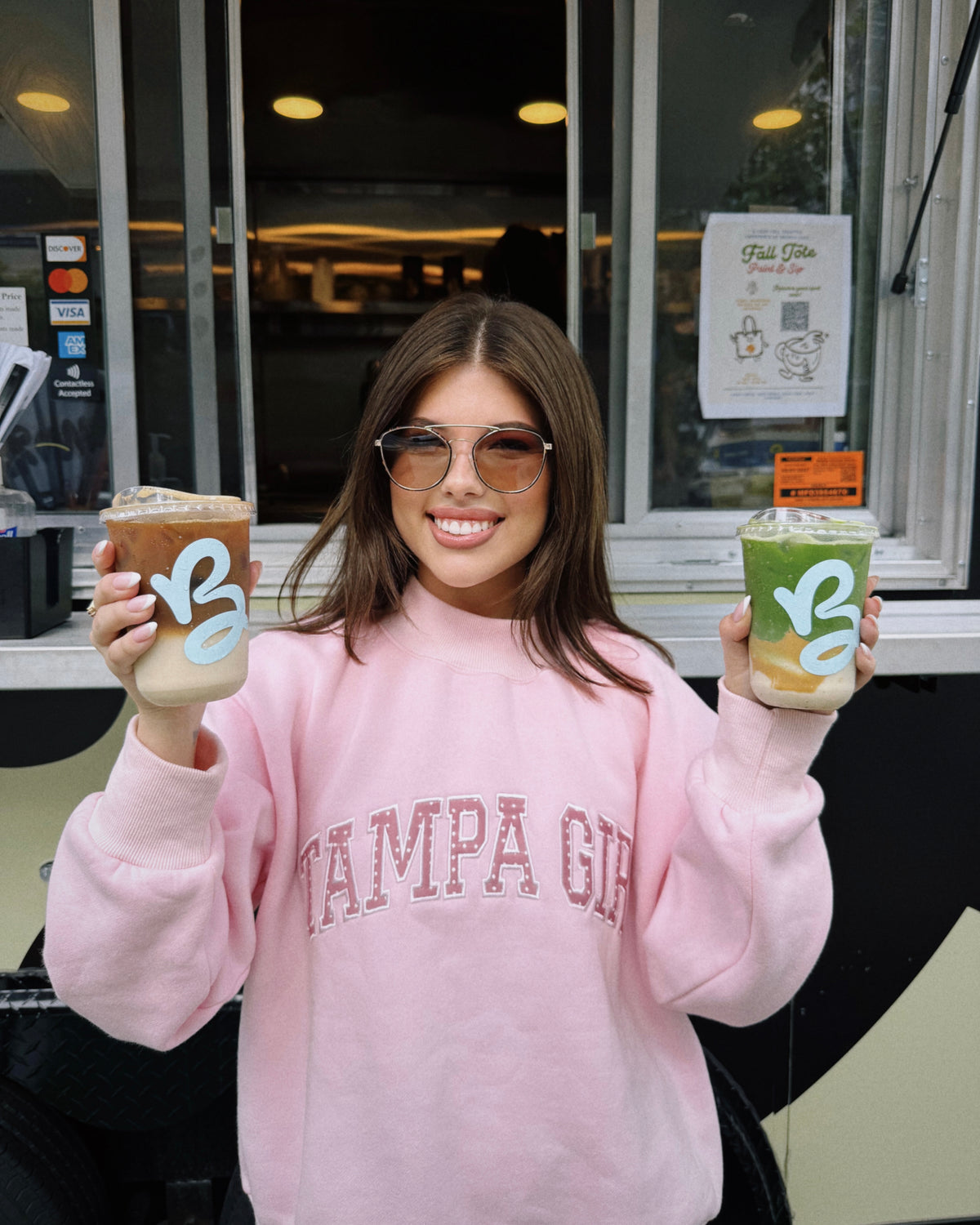 A woman wearing a pink 'Tampa Girl' sweatshirt holds up two iced coffee drinks with light blue 'VS' logos, standing in front of a cafe counter.