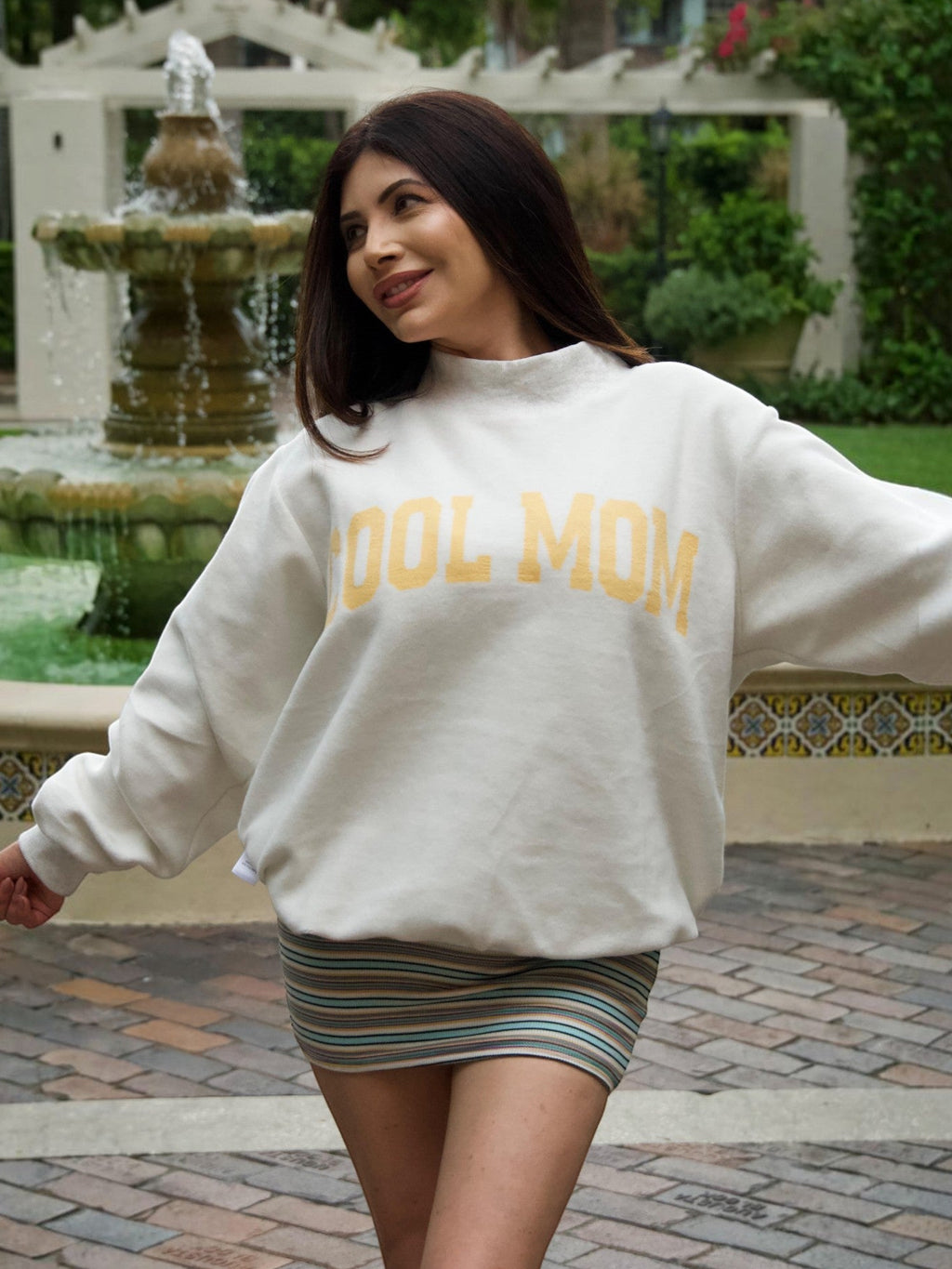 Mialur Boutique - A woman wearing an oversized cream sweatshirt with 'COOL MOM' printed in yellow across the chest, paired with a striped mini skirt, standing outdoors in front of a fountain and greenery.