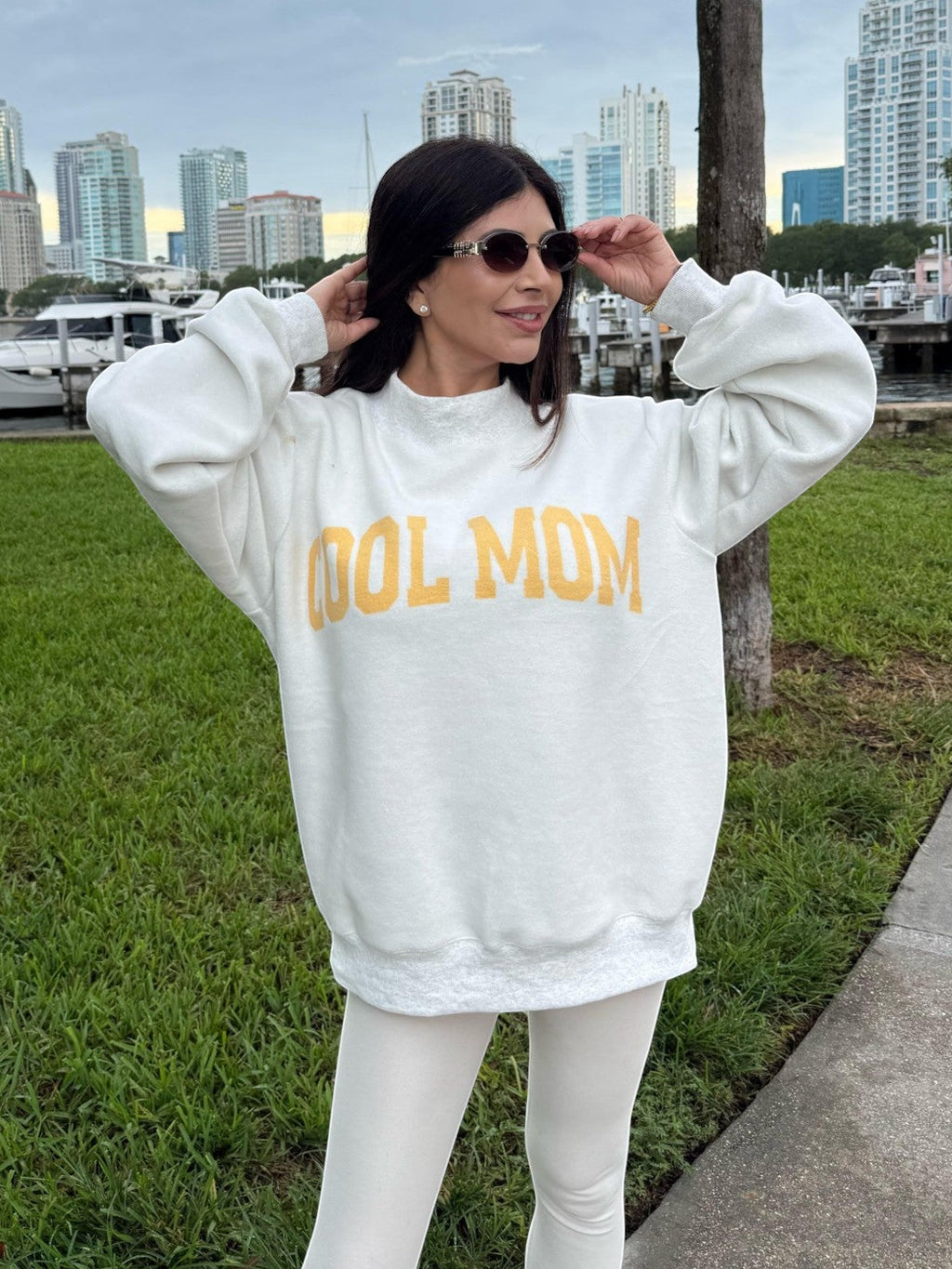 MIalur Boutique - A woman wearing a white sweatshirt with yellow 'COOL MUM' text and white leggings stands outdoors on a grassy area near a marina with a city skyline in the background.