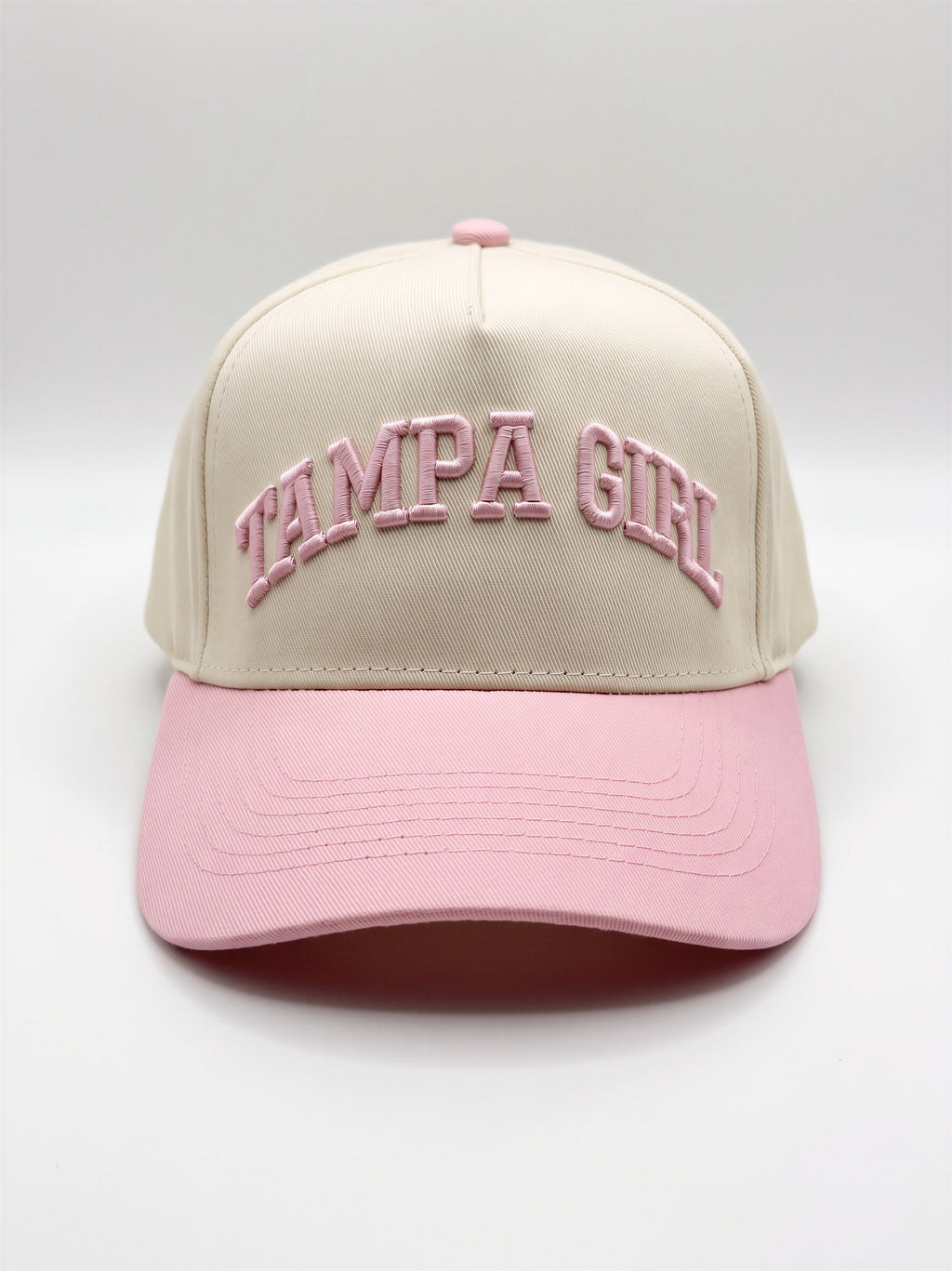 Mialur Boutique - A structured baseball cap with a cream-colored crown and a light pink brim, featuring 'TAMPA GIRL' embroidered in pink arched lettering on the front. The cap is photographed against a neutral light gray studio background.