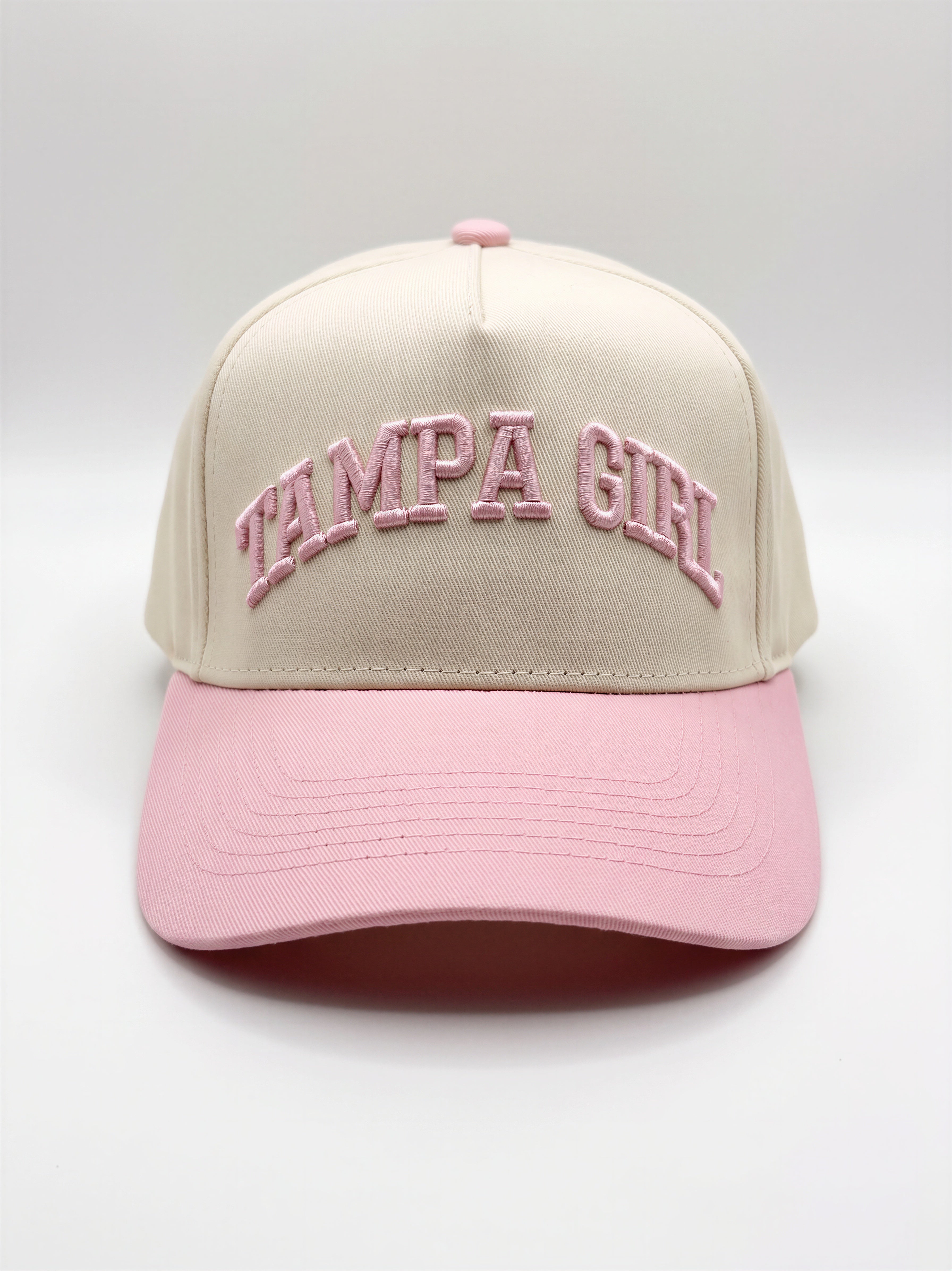 Mialur Boutique - A structured baseball cap with a cream-colored crown and a light pink brim, featuring 'TAMPA GIRL' embroidered in pink arched lettering on the front. The cap is photographed against a neutral light gray studio background.