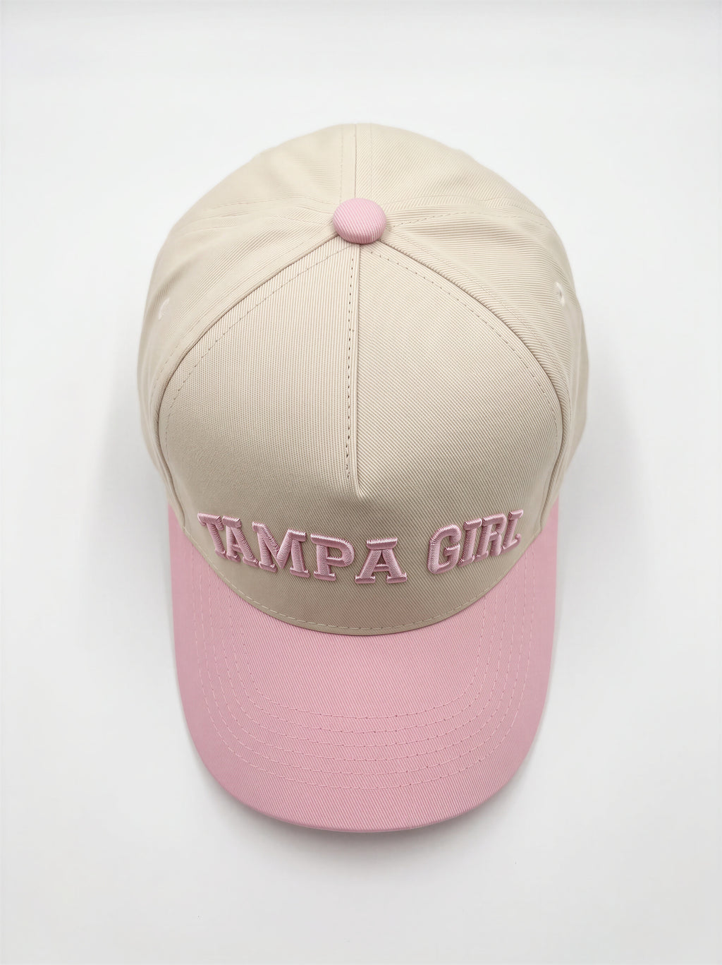 Mialur Boutique - A structured baseball cap with a beige crown and a light pink brim, featuring 'TAMPA GIRL' embroidered in pink across the front. The cap has a matching pink button on top and is photographed from a top-down angle on a clean white background.