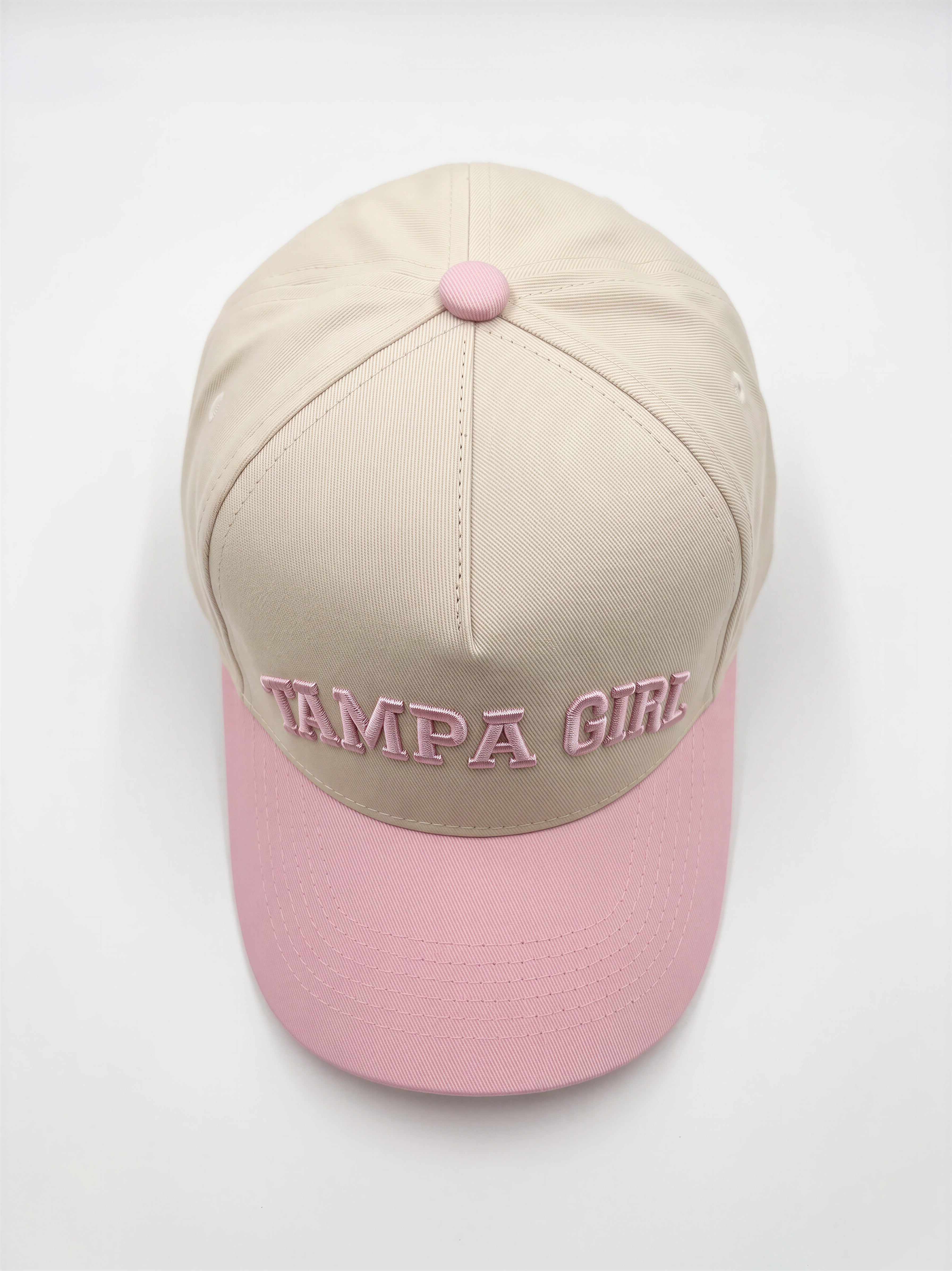 Mialur Boutique - A structured baseball cap with a beige crown and a light pink brim, featuring 'TAMPA GIRL' embroidered in pink across the front. The cap has a matching pink button on top and is photographed from a top-down angle on a clean white background.