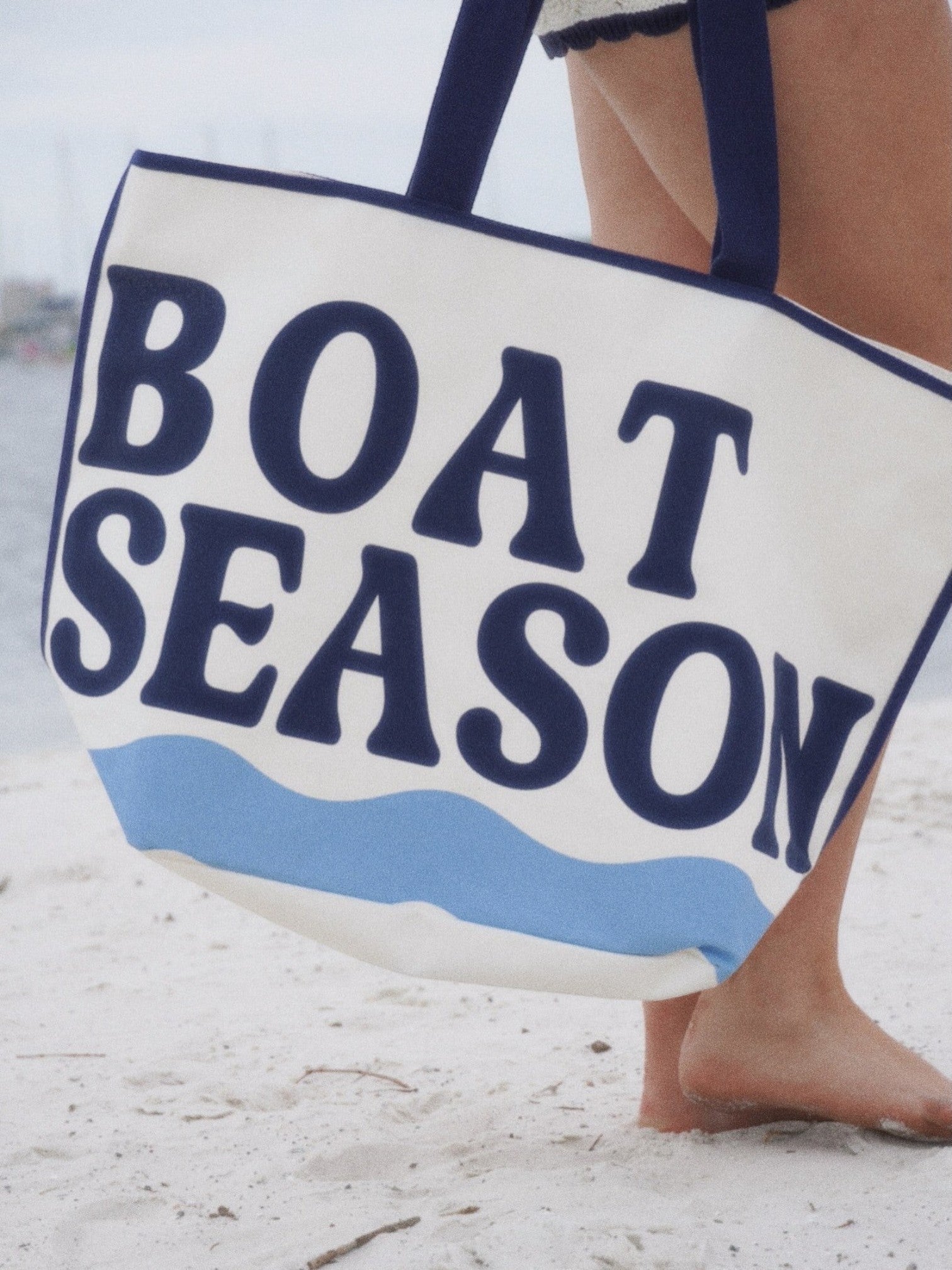 Mialur oversized ivory cotton canvas tote bag with blue BOAT SEASON lettering and wave graphic on a sandy beach