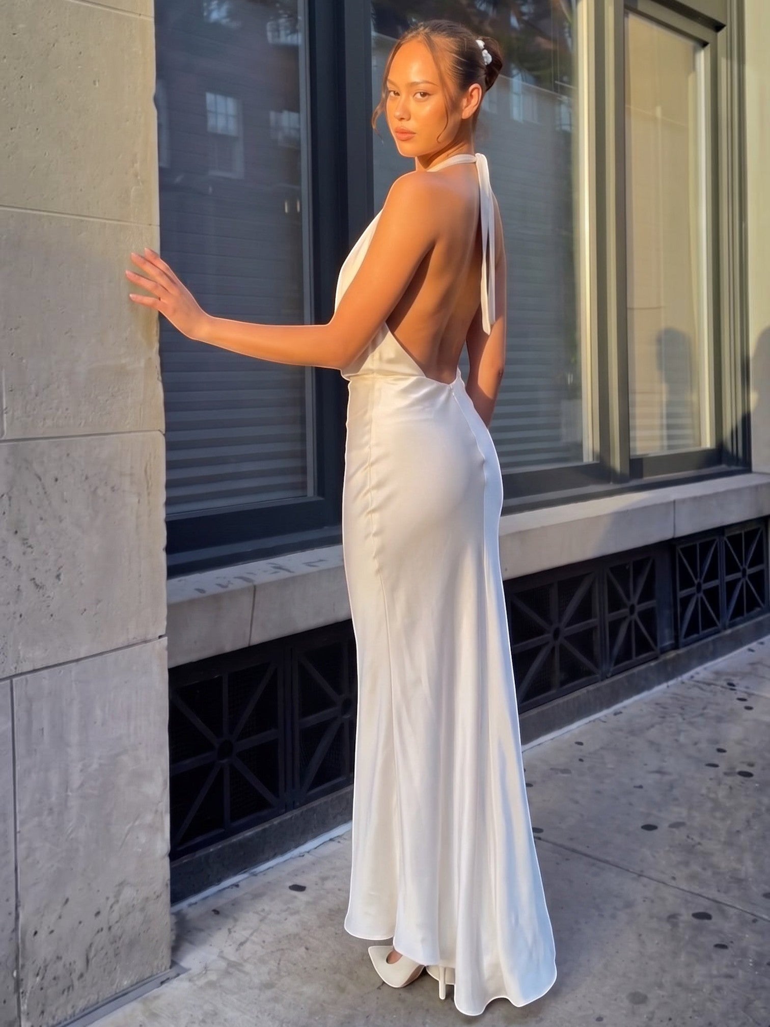 Model wearing Mialur champagne satin maxi dress with cowl neckline and open back, standing outdoors in sunlight.