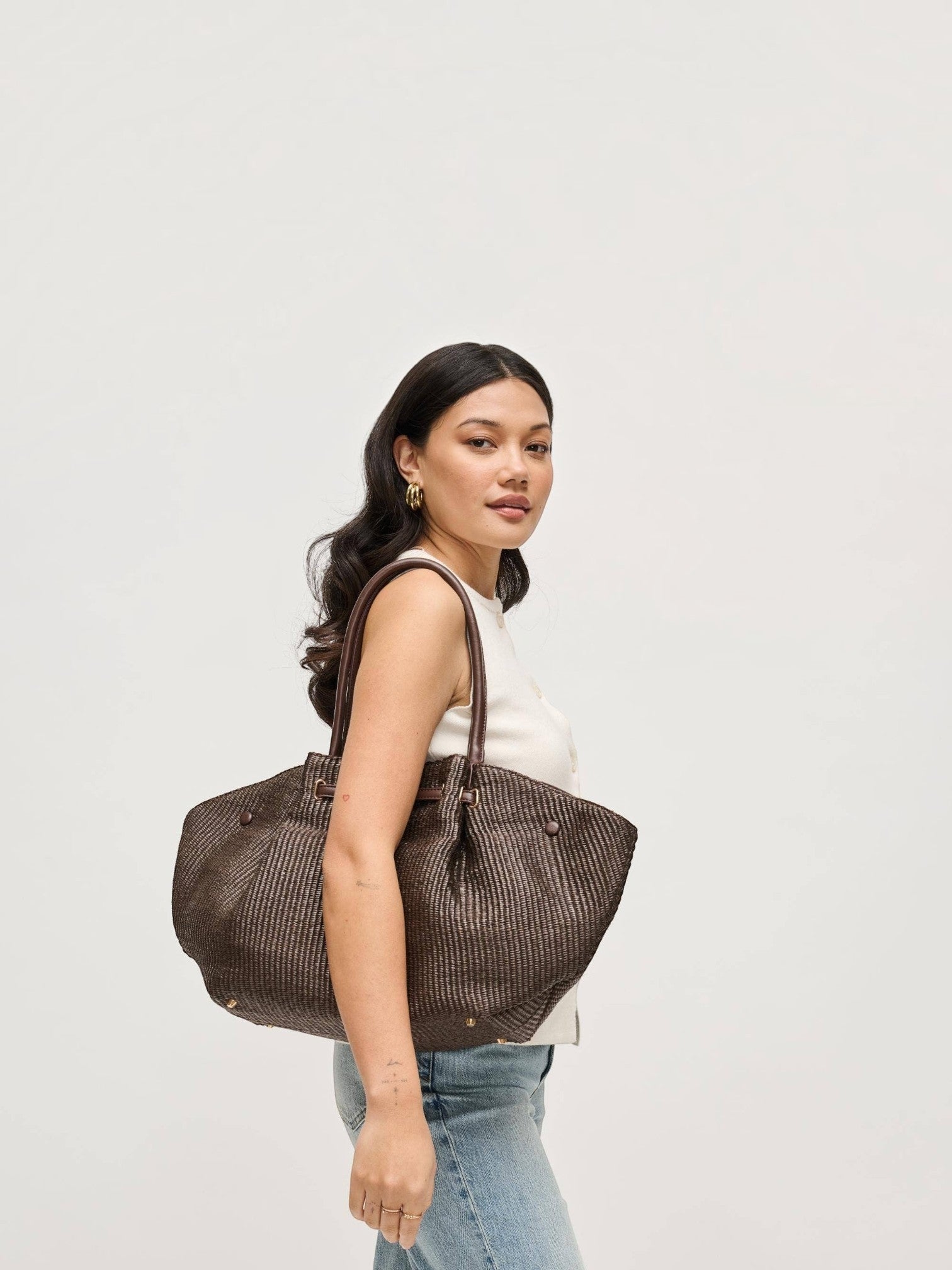 A woman wearing a Mialur chocolate brown woven tote bag with gold-tone hardware over her shoulder against a white background.