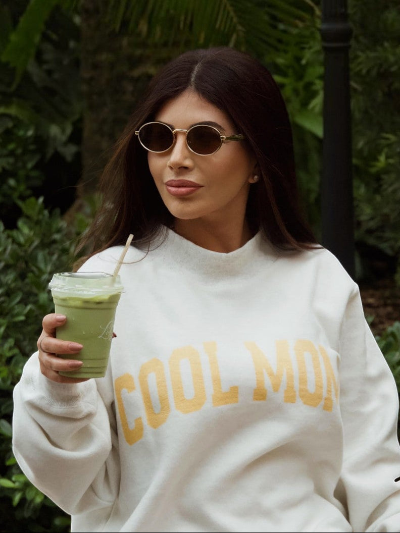 Mialur Cool Mom white mock neck sweatshirt worn by a woman holding a matcha latte in a lush garden setting.