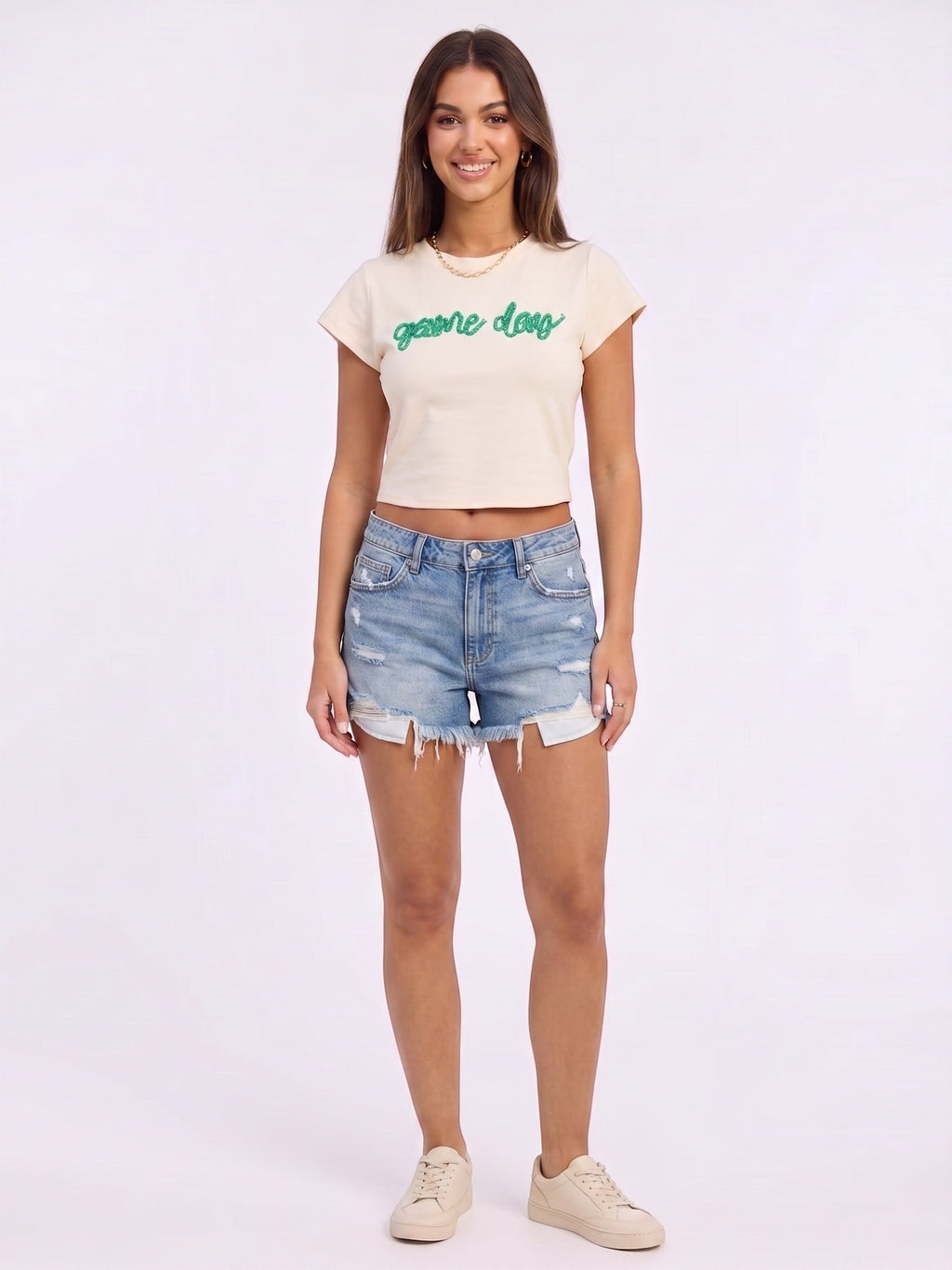 Mialur cream fitted baby tee with green game day script worn by a smiling model with denim shorts and sneakers