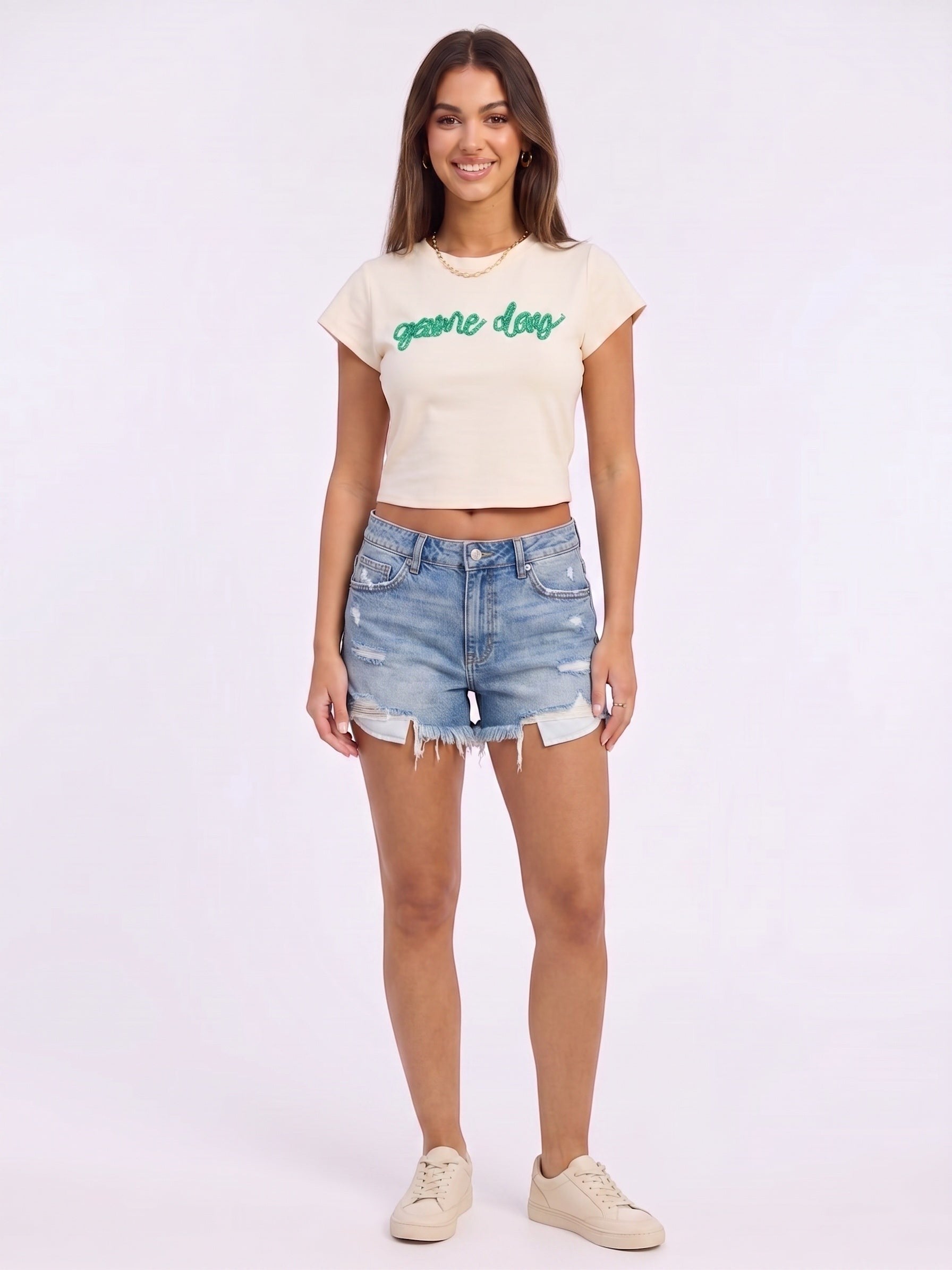 Mialur cream fitted baby tee with green game day script worn by a smiling model with denim shorts and sneakers