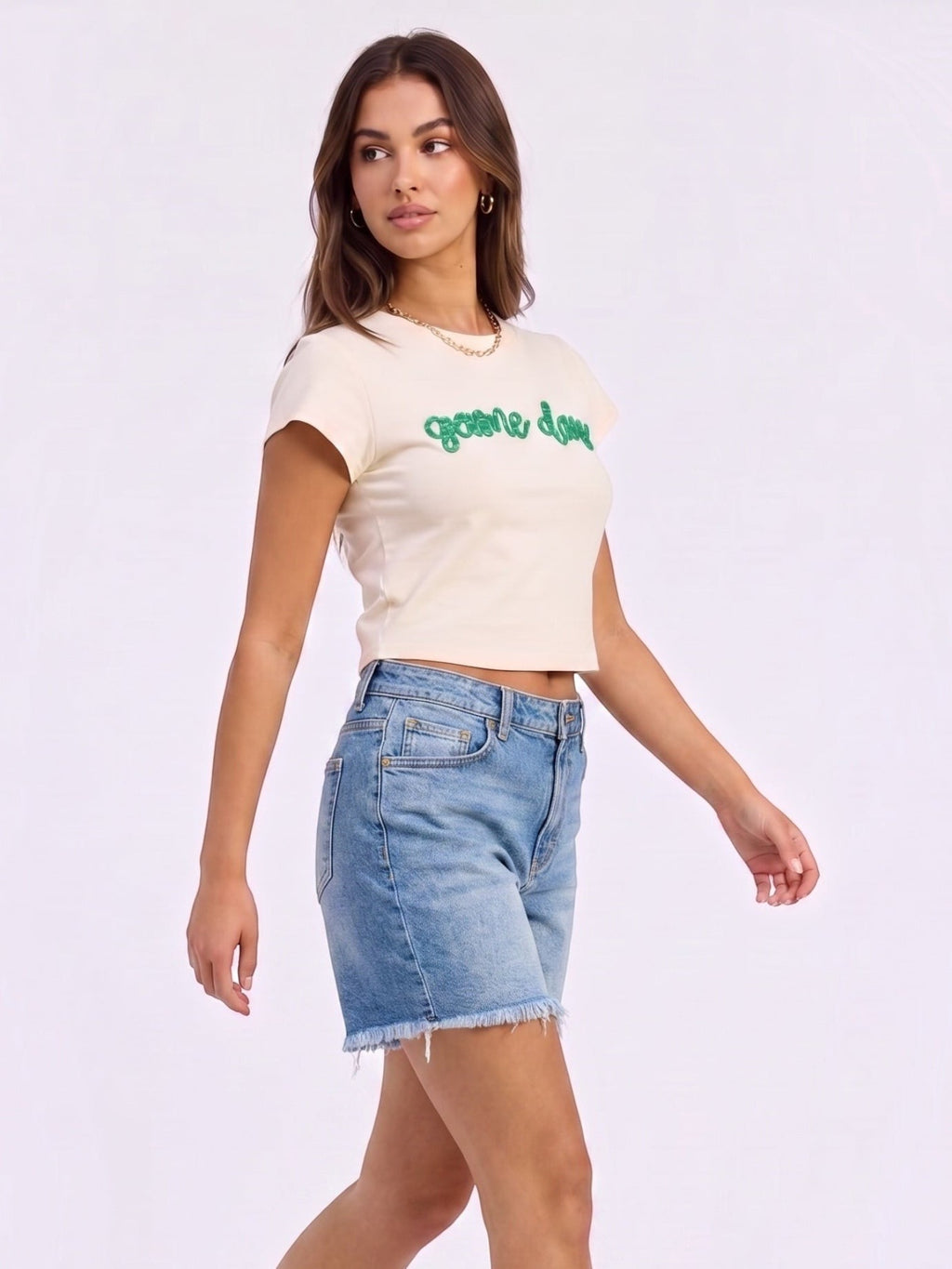 Model wearing a Mialur cream fitted baby tee with green game day script text, paired with denim shorts.