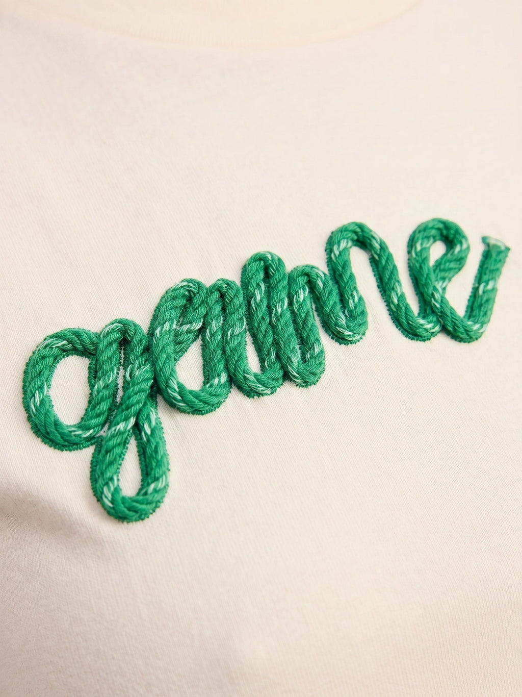Close-up of Mialur cream baby tee featuring textured green rope-style embroidery of the word game