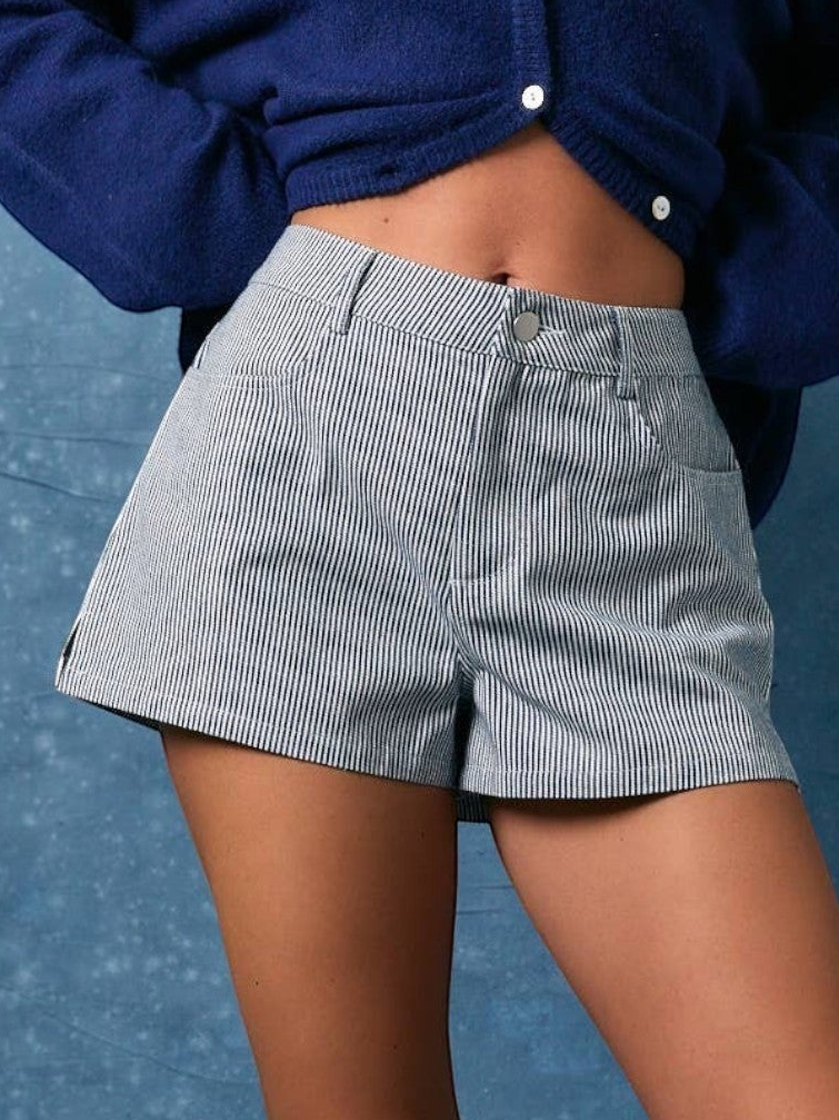 A close-up of a person wearing high-waisted, light gray pinstriped shorts with a navy blue cropped knit cardigan. The background is a textured blue wall.