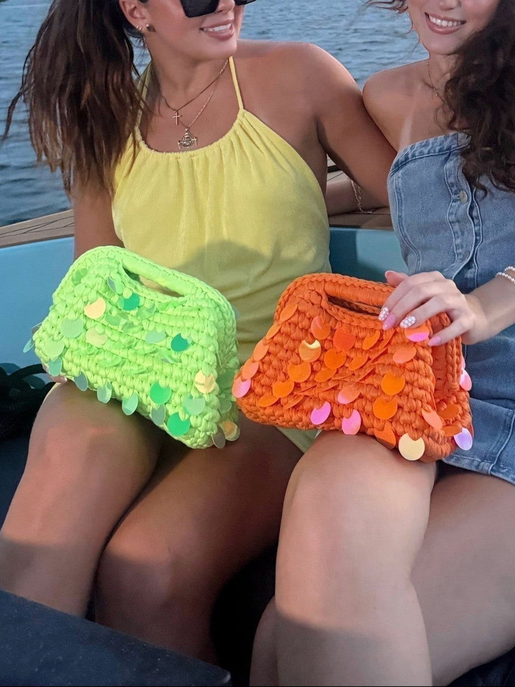 Two women on a boat holding Mialur vibrant orange and lime green sequin knit clutch bags during a summer outing.