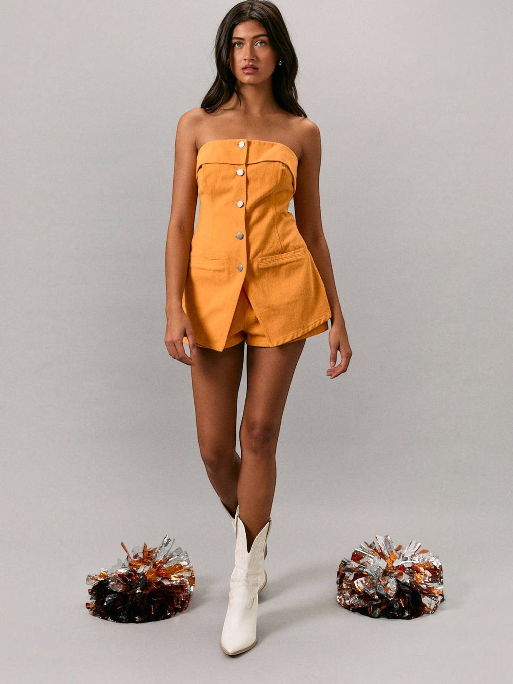 Model wearing a Mialur orange strapless cotton romper with front buttons and pockets, styled with white cowboy boots.