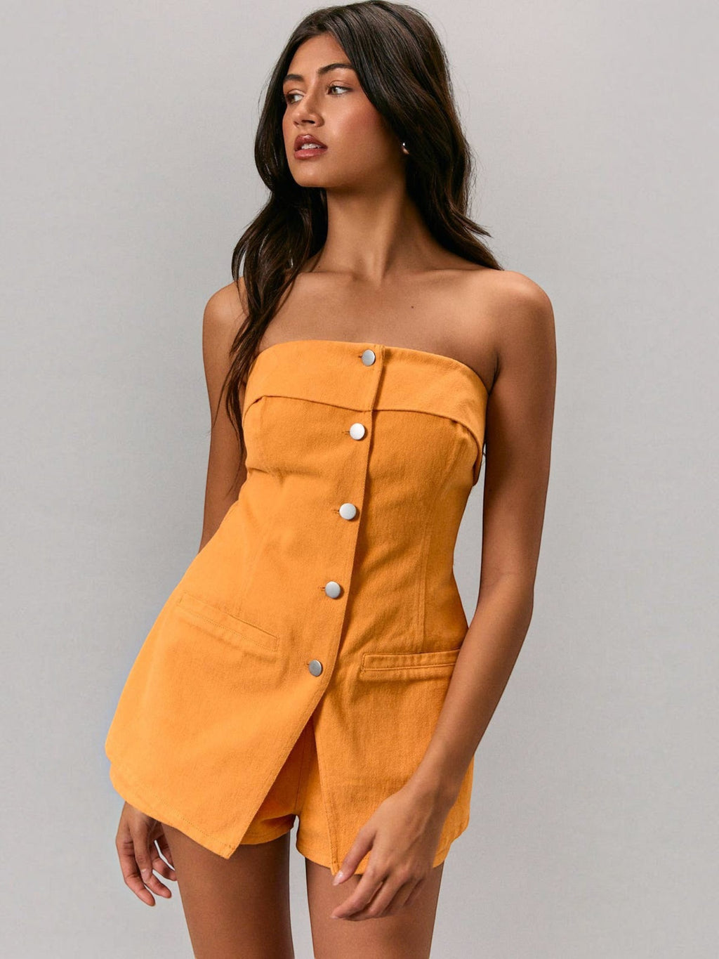 Model wearing a Mialur orange strapless cotton romper with front button closure and patch pockets against a grey background