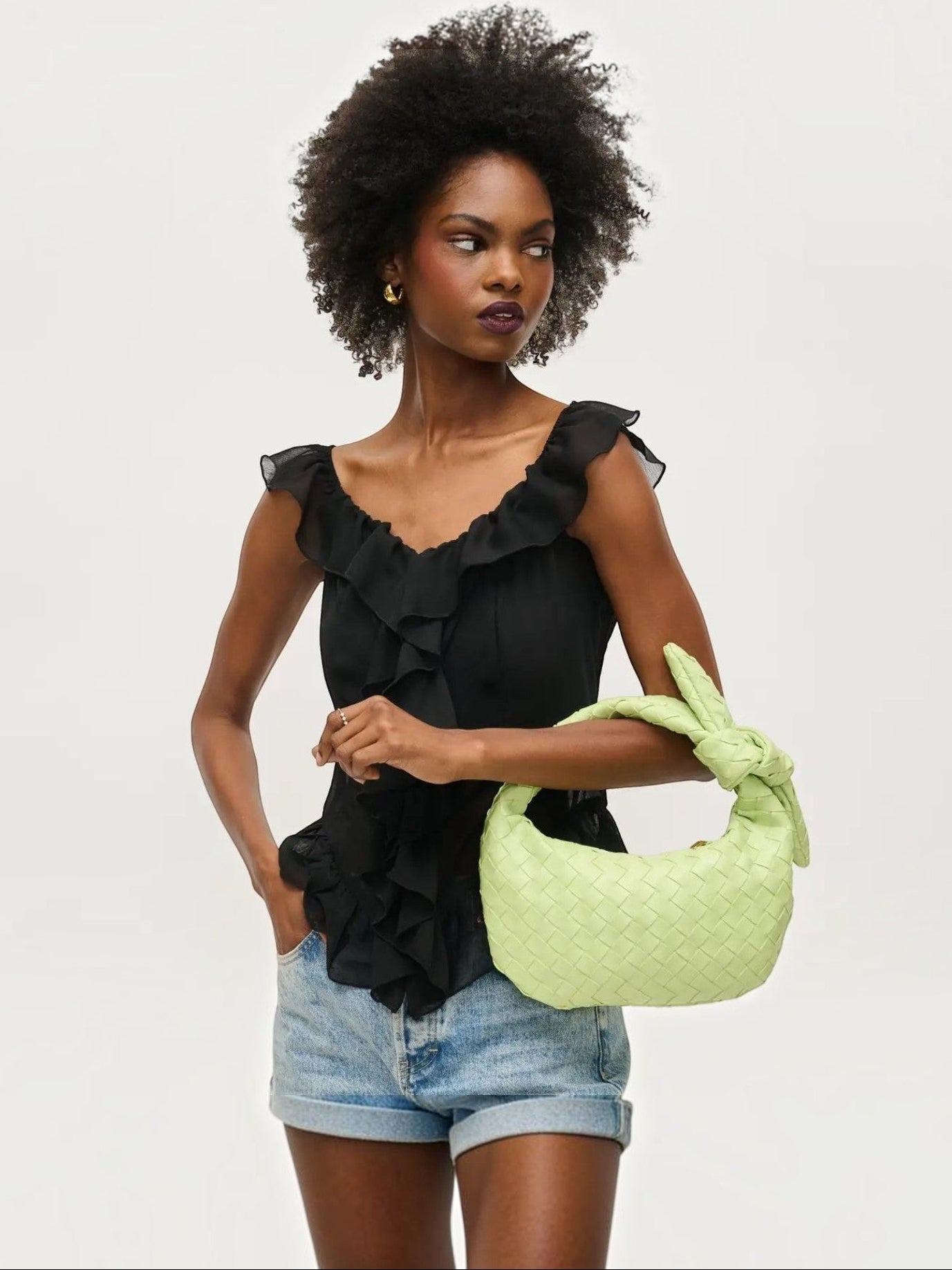 Model wearing a Mialur pistachio green woven vegan leather knot clutch with a black ruffled top and denim shorts.