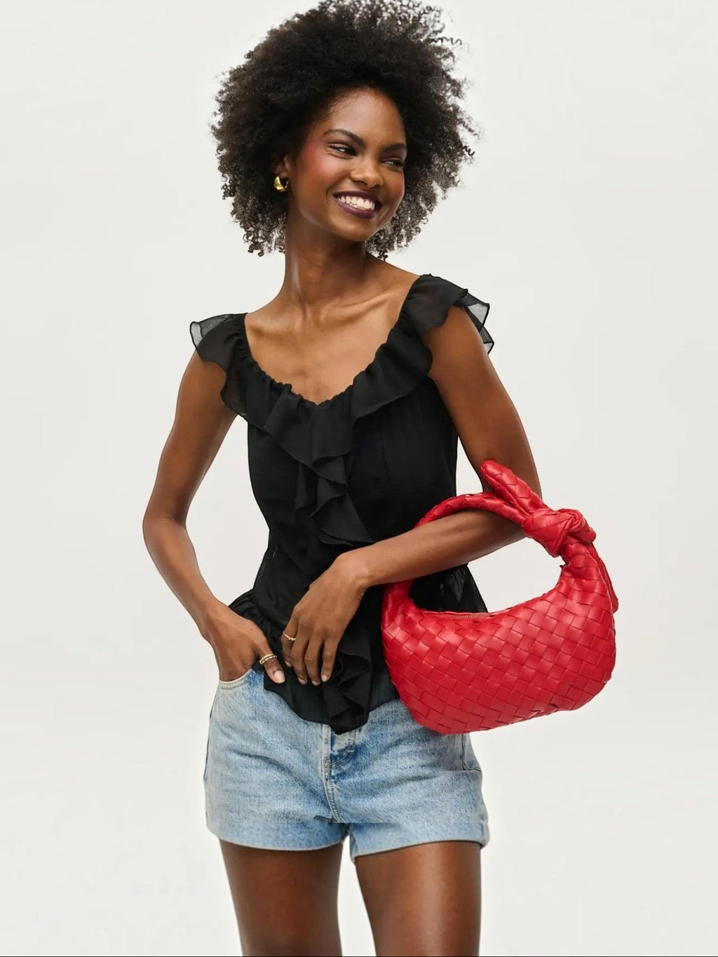 Model wearing a black ruffled top and denim shorts holding a Mialur ruby red woven vegan leather knot clutch bag.