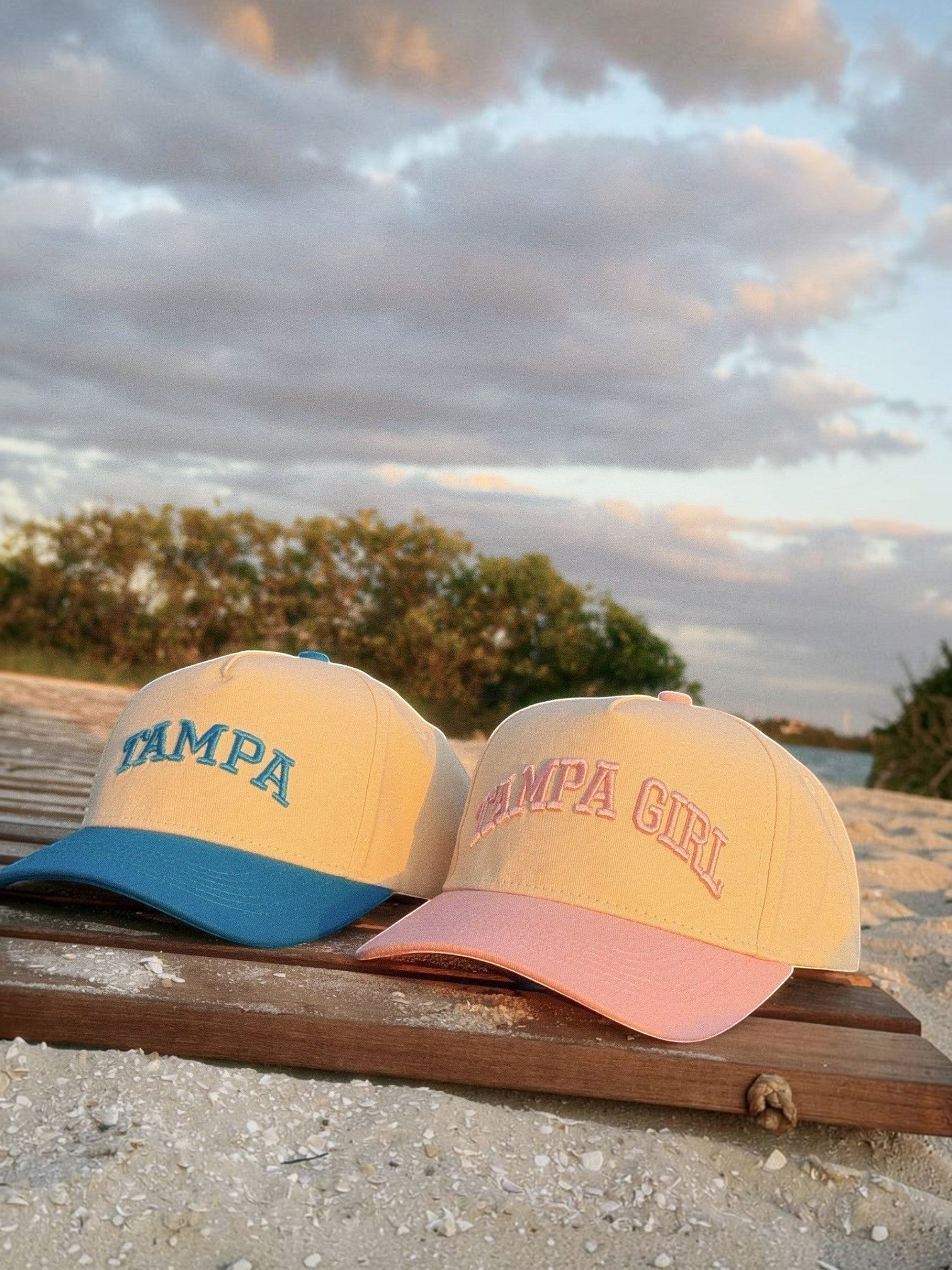 Mialur Tampa Coastal Hat in cream and blue next to a Tampa Girl hat on a wooden boardwalk at sunset.