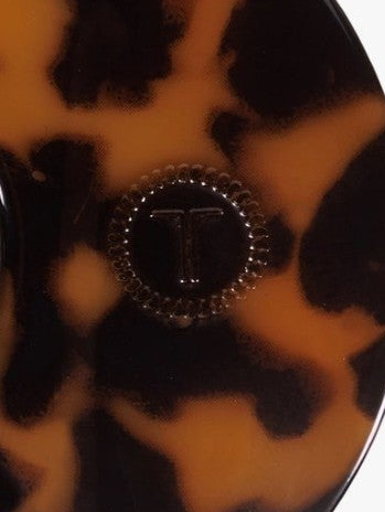 Close-up of a round tortoiseshell accessory, likely a compact mirror or case, featuring a glossy amber, black, and brown pattern with a central beaded circular detail displaying a monogram 'T'.