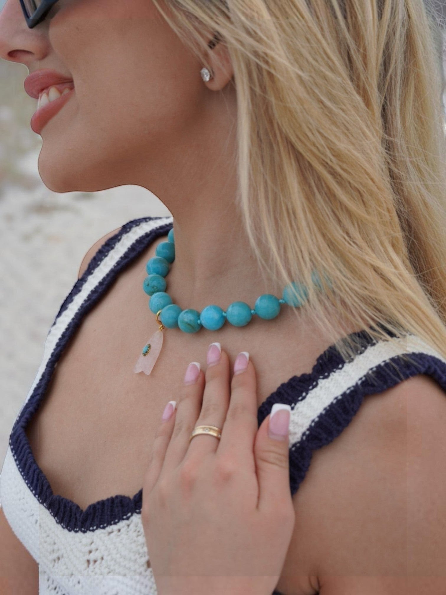 Mialur turquoise marbled bead necklace with a stone fish charm worn by a woman in a white crochet top at the beach